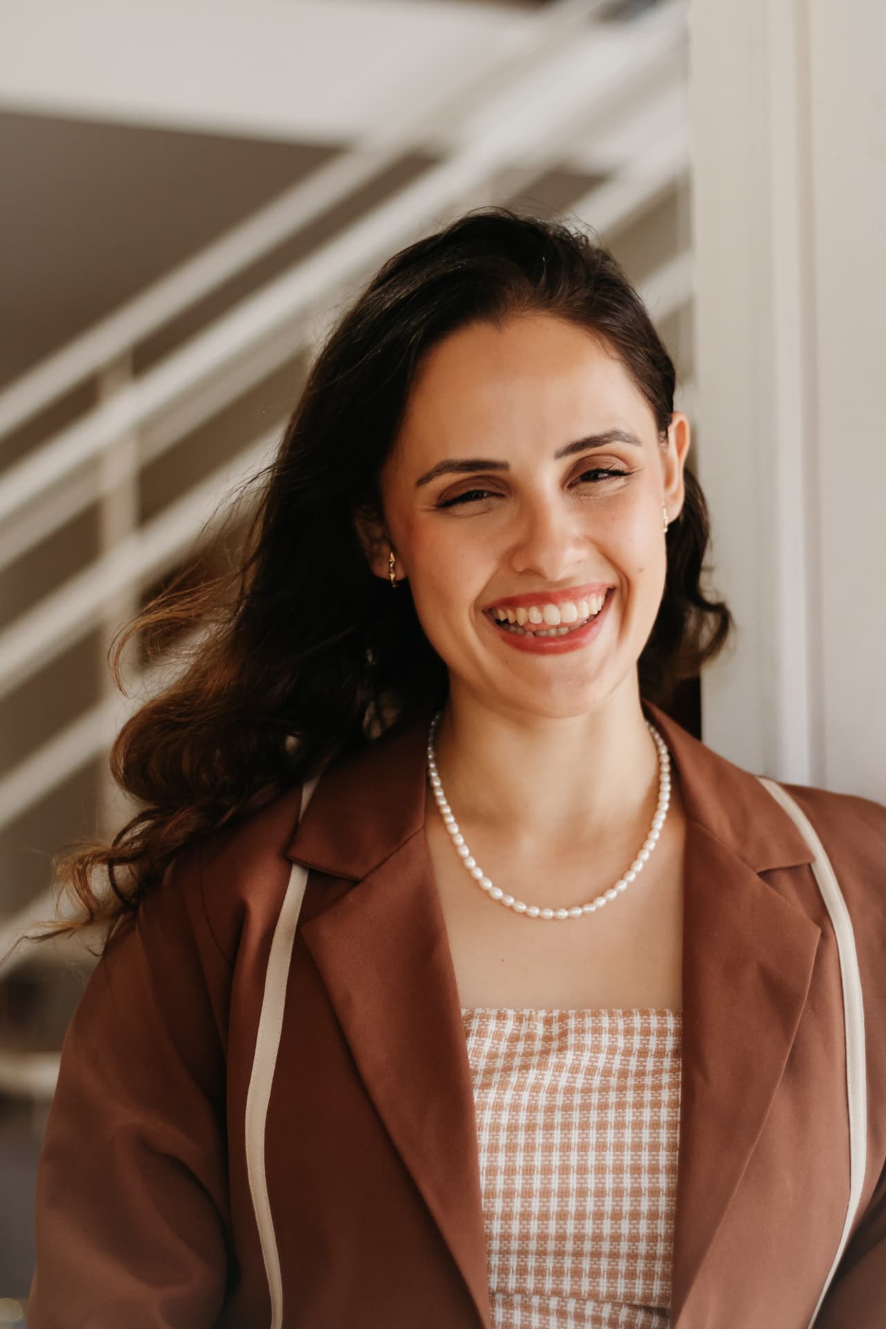 Carolina sorrindo perto da escada, com blazer marrom e colar de pérolas