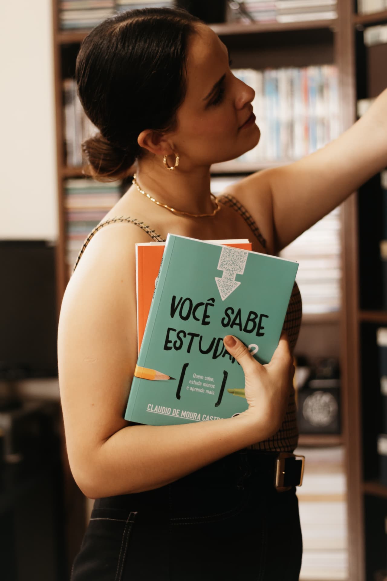 Carolina segurando livros enquanto escolhe outro na estante